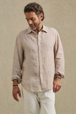 A man wears the CARUSO LINEN SHIRT by Velvet by Graham & Spencer, featuring beige and white stripes, paired with white pants as he stands facing slightly downward against a plain background.