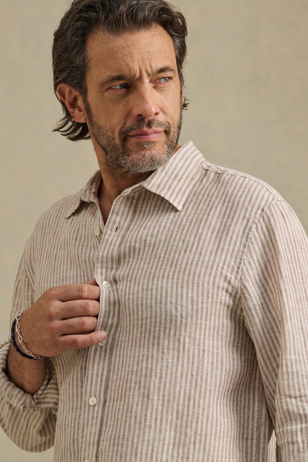 A man with short, wavy hair and a beard wears the CARUSO LINEN SHIRT by Velvet by Graham & Spencer in beige and white stripes, looking left while holding the shirt near his chest.