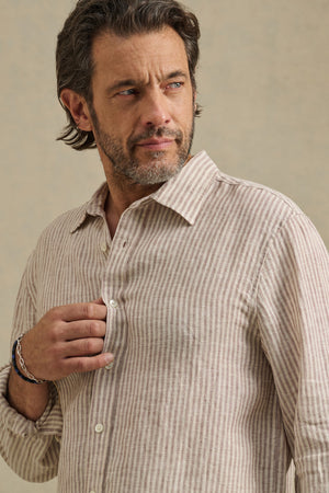 A man with short, wavy hair and a beard wears the CARUSO LINEN SHIRT by Velvet by Graham & Spencer in beige and white stripes, looking left while holding the shirt near his chest.