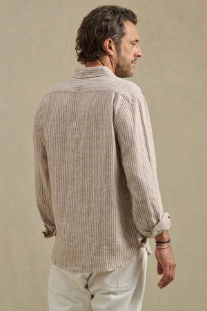 A man stands facing away, wearing the CARUSO LINEN SHIRT by Velvet by Graham & Spencer—a beige and white striped long-sleeve shirt—paired with white pants against a neutral background.