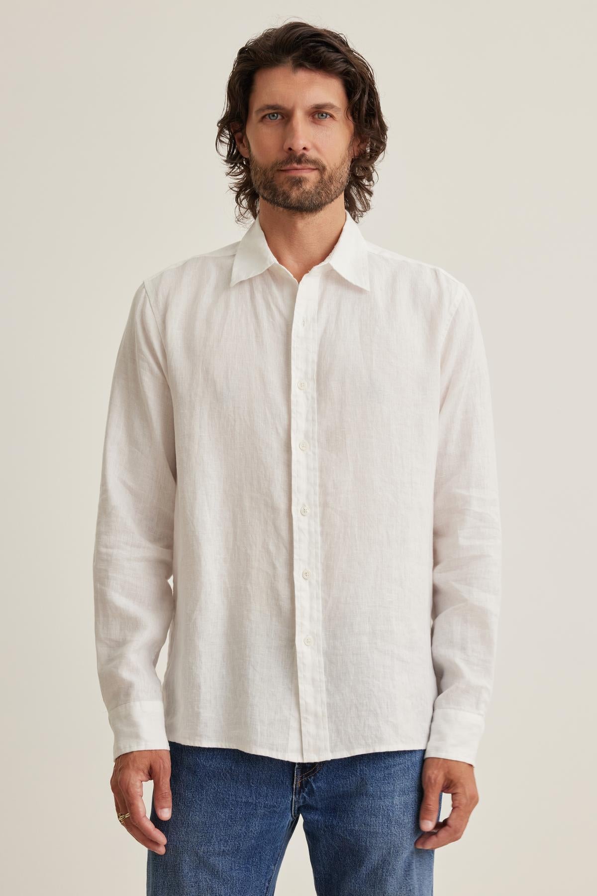 A man with wavy hair wears a CARUSO LINEN BUTTON-UP SHIRT by Velvet by Graham & Spencer and blue jeans, standing against a plain background.-38643242893505