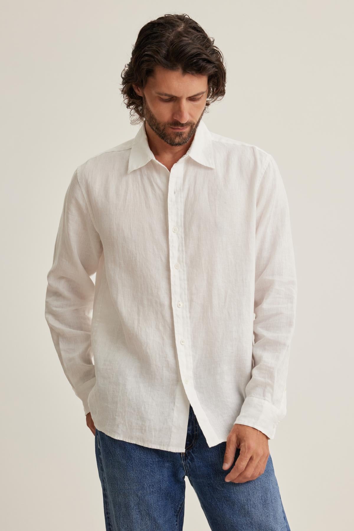   A man with medium-length hair wears a CARUSO LINEN BUTTON-UP SHIRT from Velvet by Graham & Spencer paired with blue jeans. He gazes downward with a neutral expression against a plain background. 