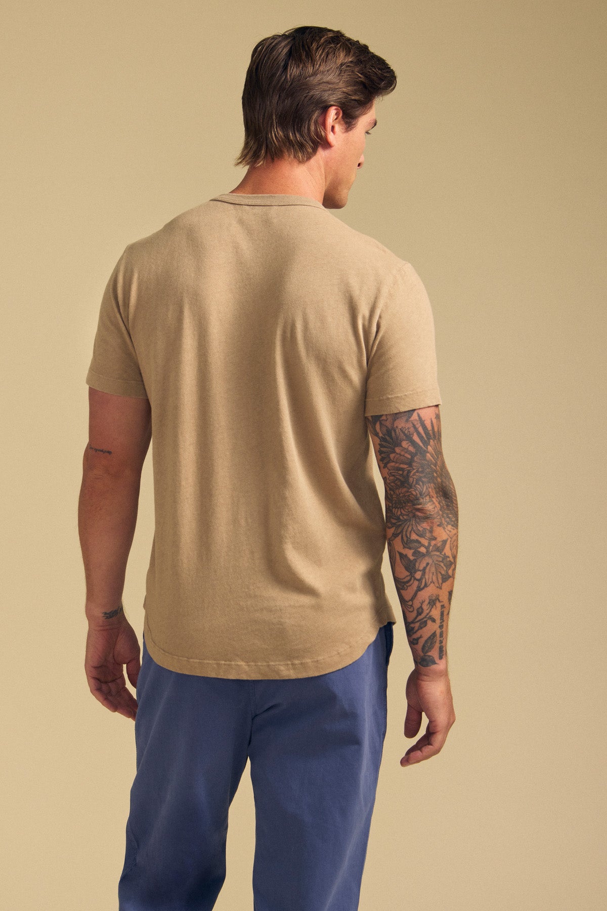A man faces away wearing the LUKA TEE by Velvet by Graham & Spencer—a beige crew neck made from lightweight cotton-linen—paired with blue pants. A dark floral tattoo covers his right arm, set against a plain light tan backdrop for warm-weather style.