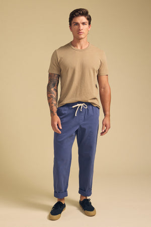 A man stands against a plain beige background wearing the LUKA TEE by Velvet by Graham & Spencer, a beige crew neck t-shirt in lightweight cotton-linen, paired with blue drawstring pants and black sneakers.