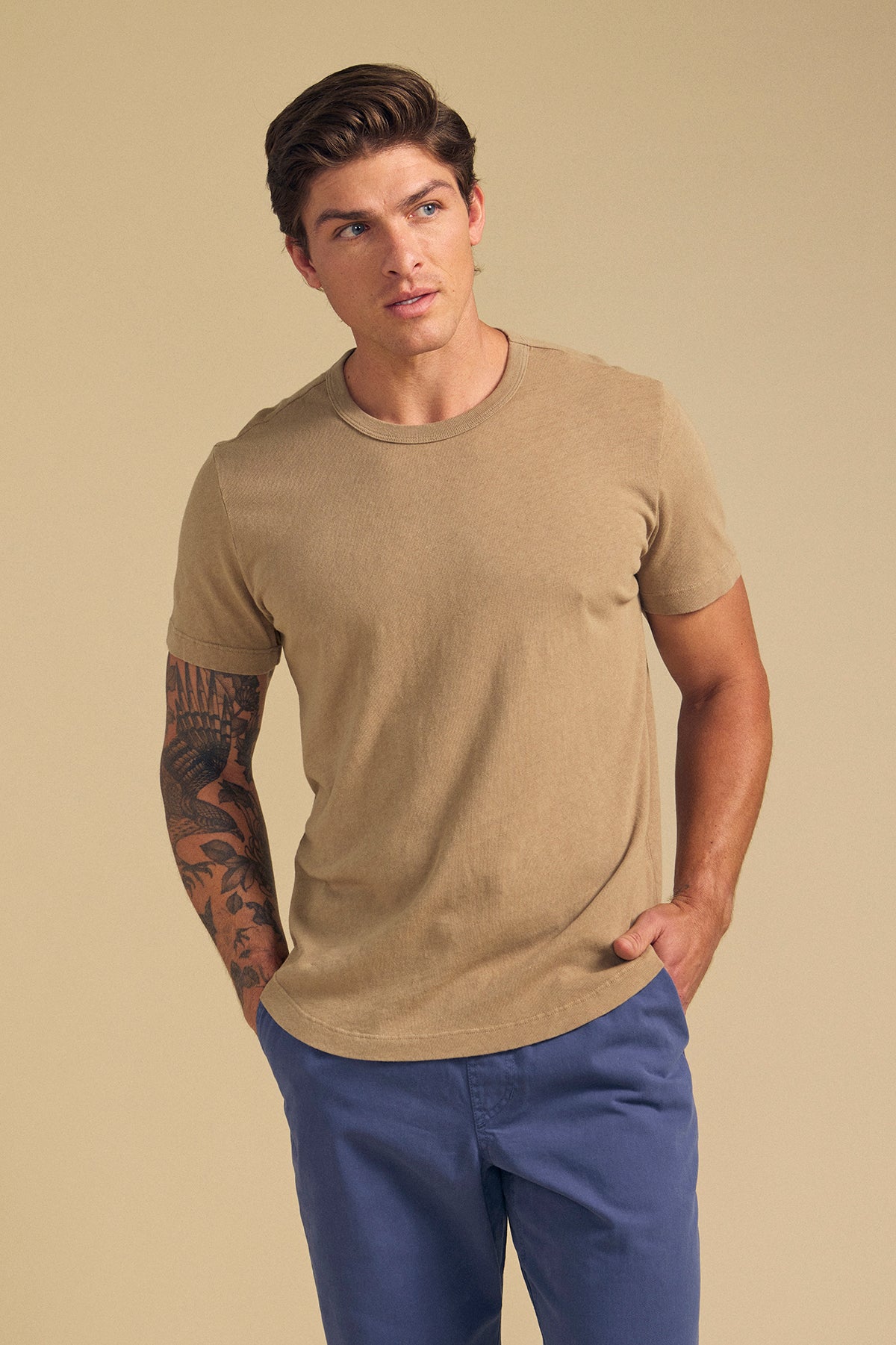 A man with short brown hair wears the LUKA TEE by Velvet by Graham & Spencer—a beige crew neck in lightweight cotton-linen—paired with blue pants, standing against a plain beige background; ideal for warm-weather days.-41617466589377
