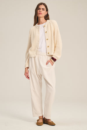 A woman wears the Velvet by Graham & Spencer LANEY CORDUROY PANT in cream with a knit cardigan, white shirt, and brown flats, standing against a plain light background.