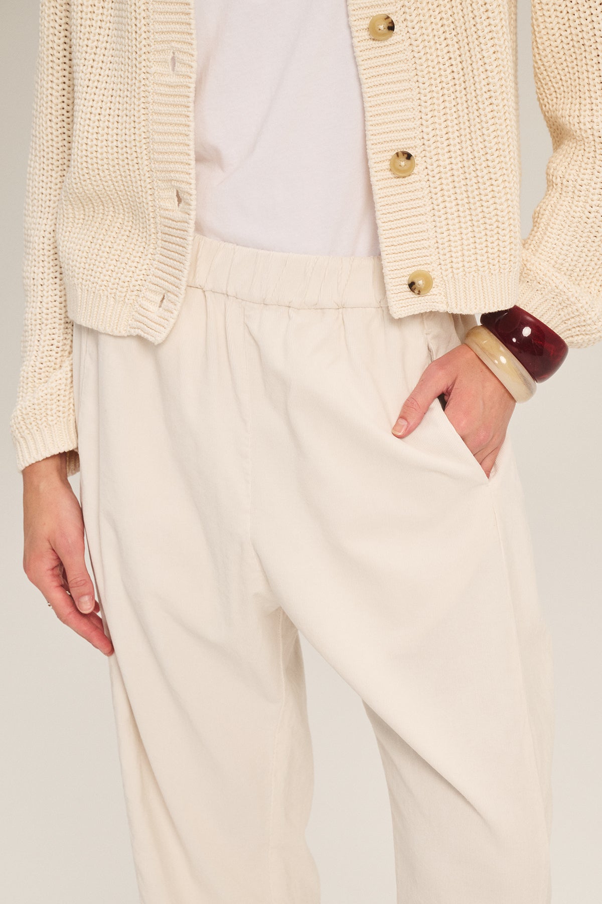 A person wears the Velvet by Graham & Spencer LANEY CORDUROY PANT in cream with a coordinating cardigan, white t-shirt, chunky bangles, and one hand in their pocket.-39574404104385