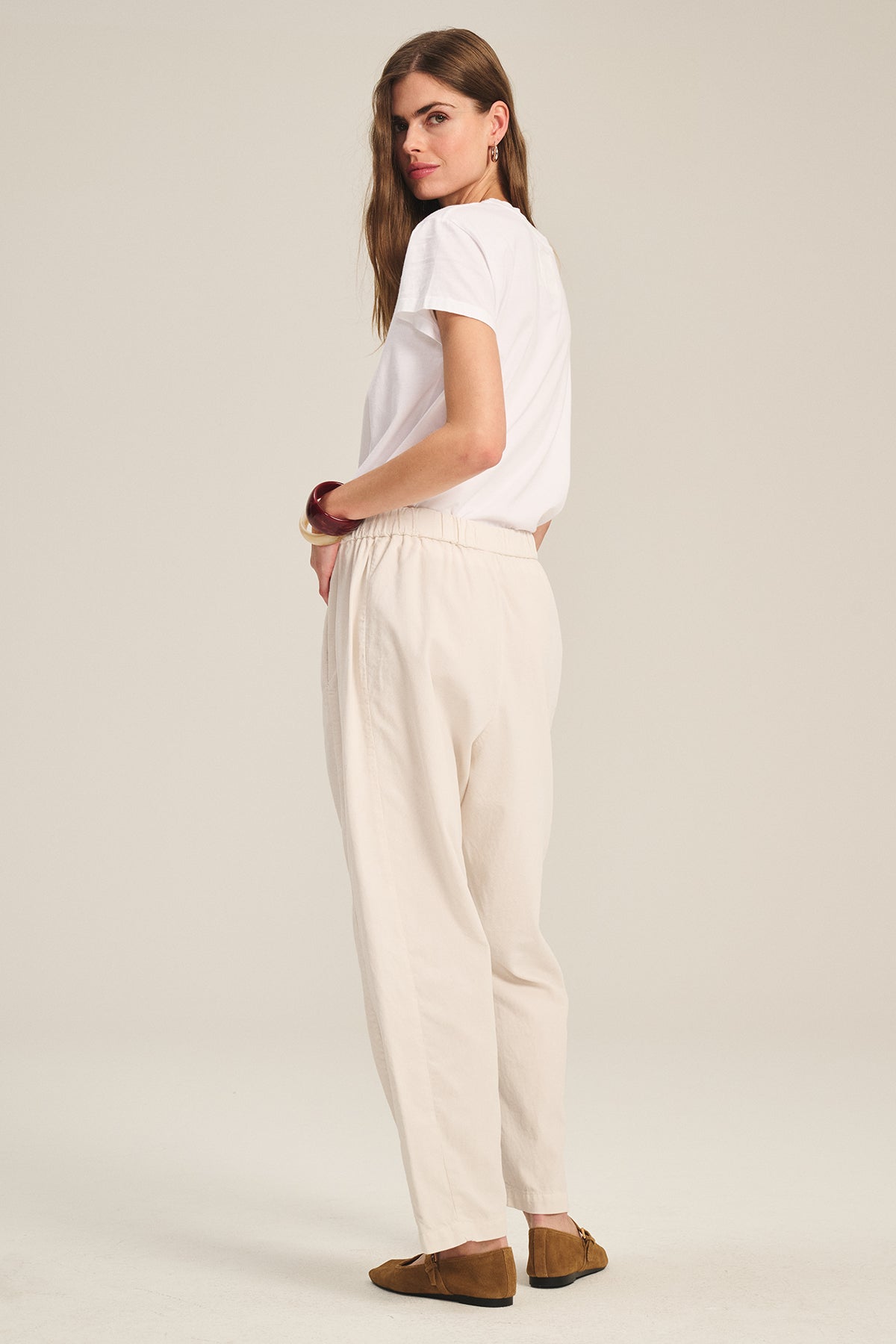 A woman stands sideways, looking at the camera, wearing Velvet by Graham & Spencer's LANEY CORDUROY PANT in light beige with a white T-shirt and brown shoes, set against a plain light background.-39574404137153