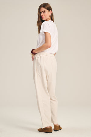 A woman stands sideways, looking at the camera, wearing Velvet by Graham & Spencer's LANEY CORDUROY PANT in light beige with a white T-shirt and brown shoes, set against a plain light background.