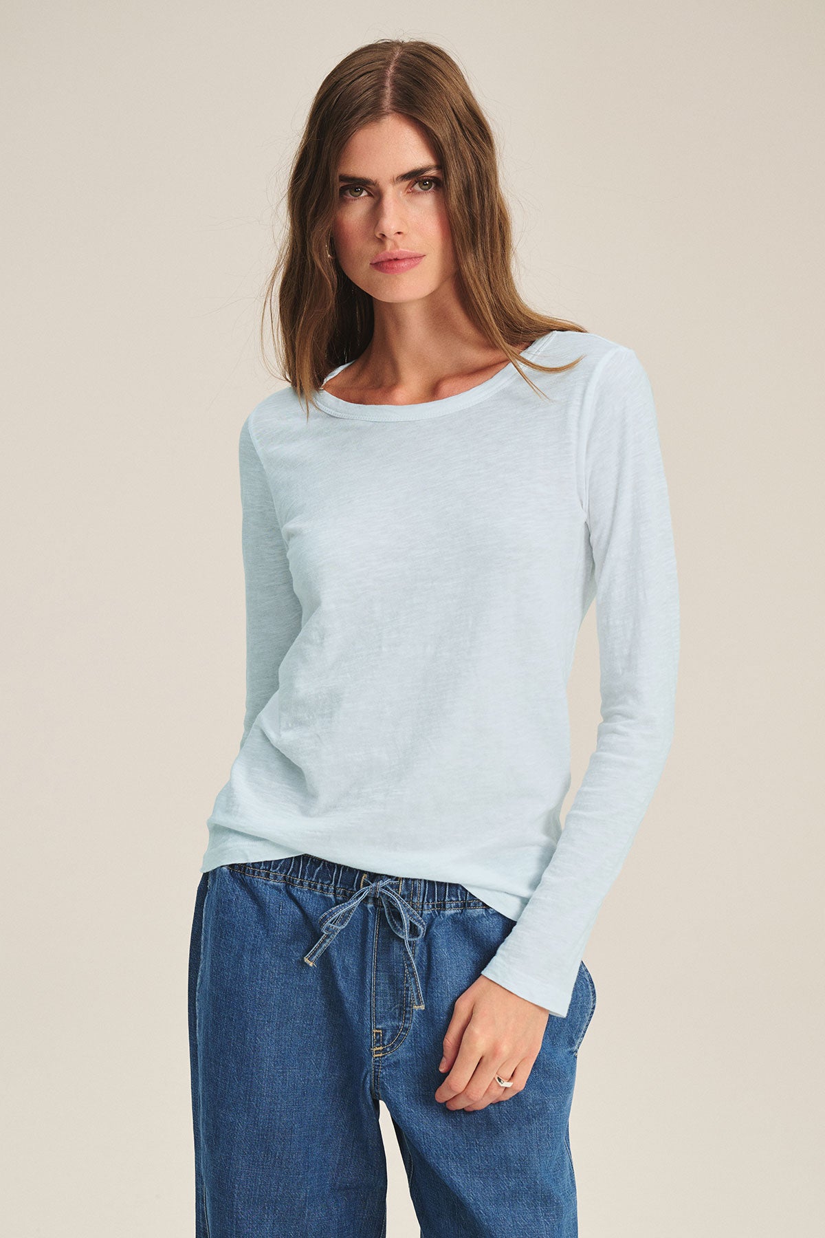 A woman wears the LIZZIE TEE by Velvet by Graham & Spencer—a light blue long-sleeve crewneck—paired with blue drawstring pants, standing against a plain background.-40416944685249