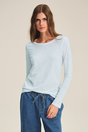 A woman wears the LIZZIE TEE by Velvet by Graham & Spencer—a light blue long-sleeve crewneck—paired with blue drawstring pants, standing against a plain background.