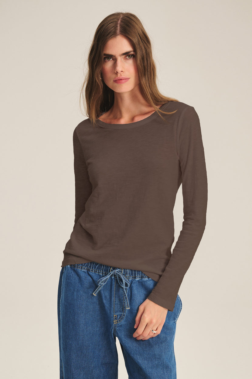 A woman wears the LIZZIE TEE by Velvet by Graham & Spencer, a brown long sleeve crew neck top, paired with blue drawstring pants, standing against a plain light background.