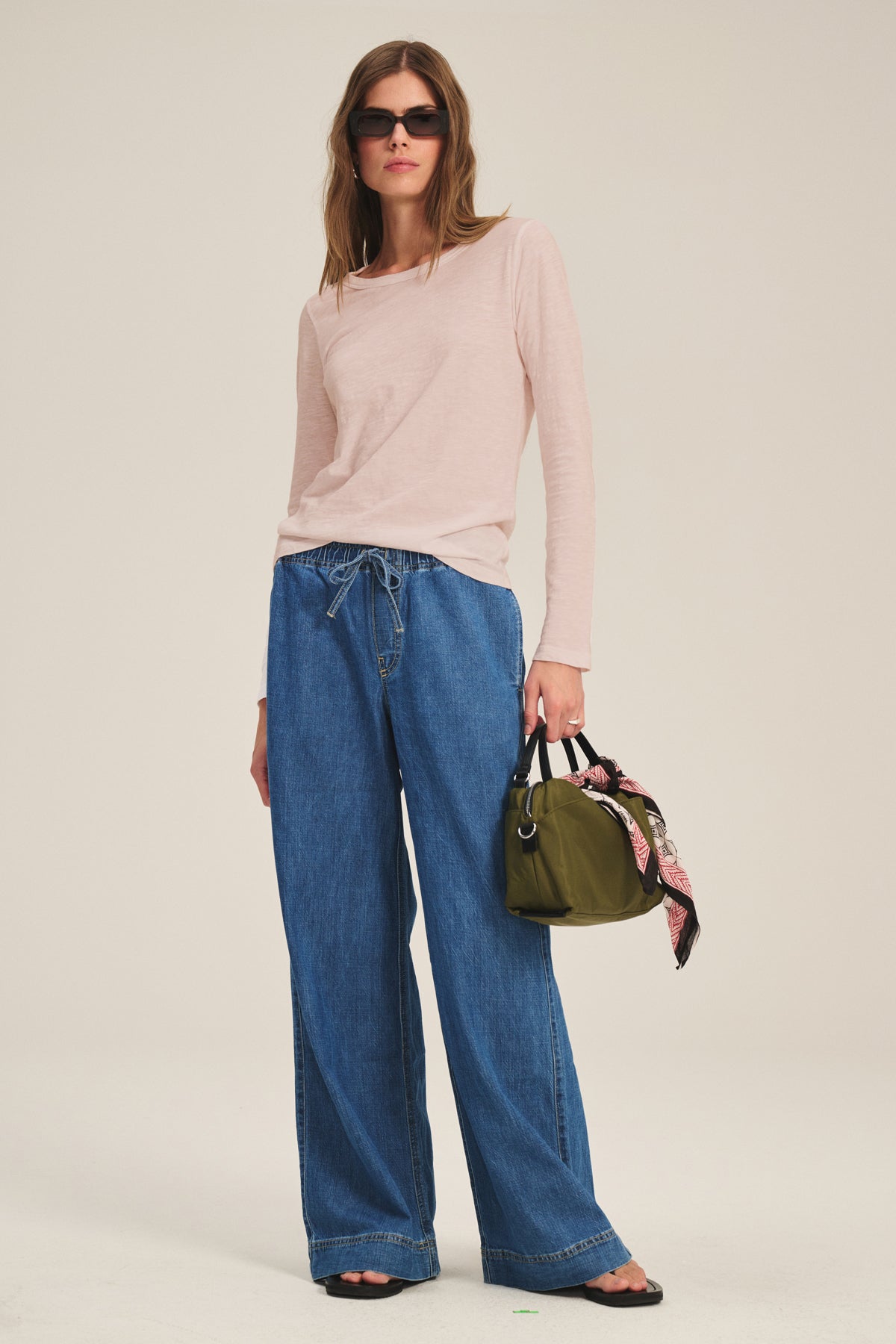 A person wearing sunglasses, the LIZZIE TEE by Velvet by Graham & Spencer in light pink, wide-leg blue jeans, sandals, and holding an olive green handbag with a printed scarf tied to it.-40163417653441