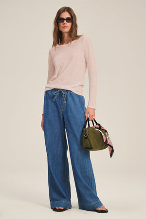 A person wearing sunglasses, the LIZZIE TEE by Velvet by Graham & Spencer in light pink, wide-leg blue jeans, sandals, and holding an olive green handbag with a printed scarf tied to it.