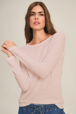 A woman with long brown hair models the LIZZIE TEE by Velvet by Graham & Spencer, a light pink long-sleeve top, paired with blue pants against a plain beige background.