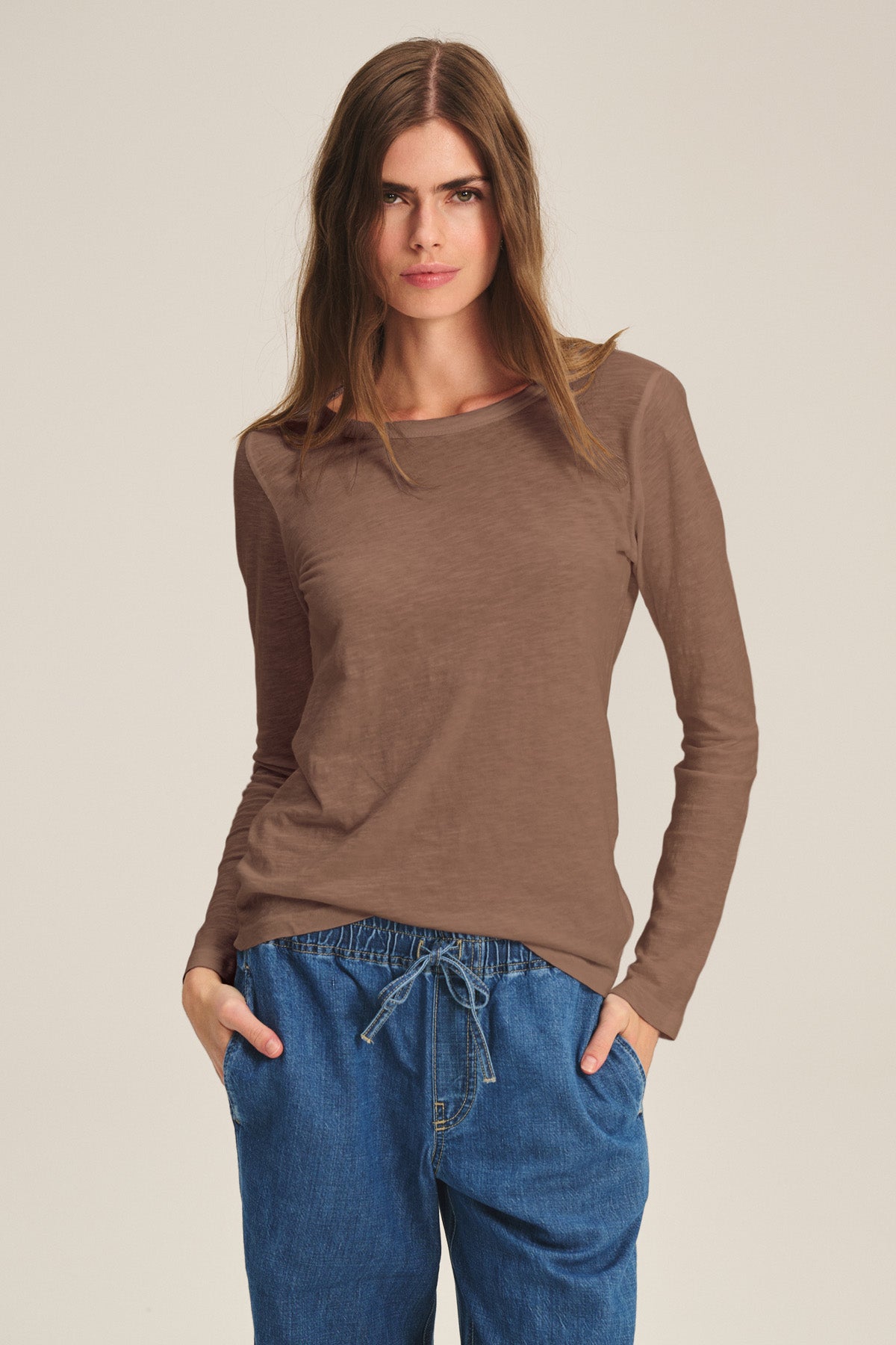 A woman stands with hands in pockets, wearing the LIZZIE TEE by Velvet by Graham & Spencer—a brown long-sleeve cotton slub crewneck tee—paired with blue drawstring pants against a plain light background.-40921275793601