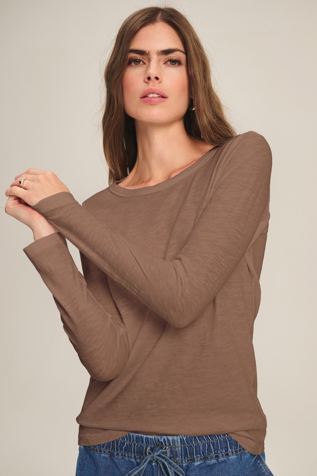 A woman with long brown hair models the LIZZIE TEE by Velvet by Graham & Spencer—a cotton slub crewneck with long sleeves—paired with blue drawstring pants, set against a plain light background.-40921275826369