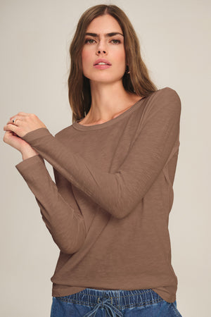 A woman with long brown hair models the LIZZIE TEE by Velvet by Graham & Spencer—a cotton slub crewneck with long sleeves—paired with blue drawstring pants, set against a plain light background.
