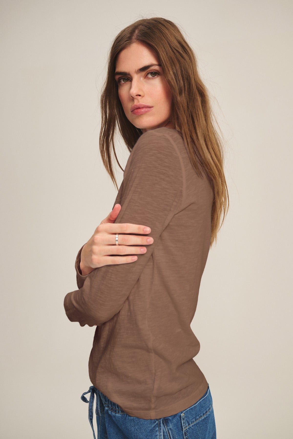 A woman with long brown hair wears the LIZZIE TEE by Velvet by Graham & Spencer—a brown cotton slub crew neck—paired with blue jeans, standing sideways and looking at the camera with a neutral expression.-40921275859137