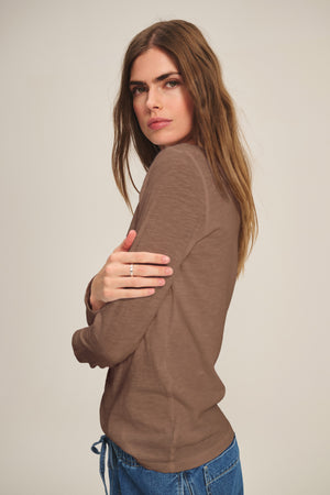 A woman with long brown hair wears the LIZZIE TEE by Velvet by Graham & Spencer—a brown cotton slub crew neck—paired with blue jeans, standing sideways and looking at the camera with a neutral expression.