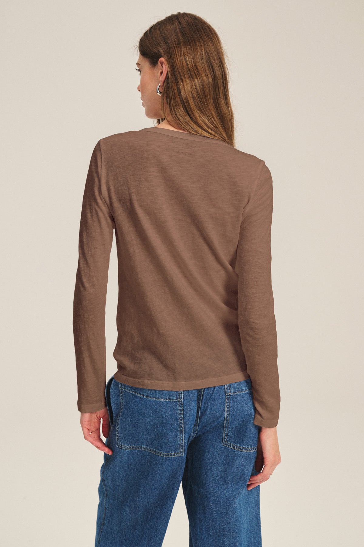 A woman faces away, wearing the LIZZIE TEE by Velvet by Graham & Spencer—a long-sleeve cotton slub top with a crew neck—paired with blue jeans, set against a plain light background.