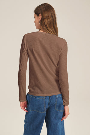 A woman faces away, wearing the LIZZIE TEE by Velvet by Graham & Spencer—a long-sleeve cotton slub top with a crew neck—paired with blue jeans, set against a plain light background.