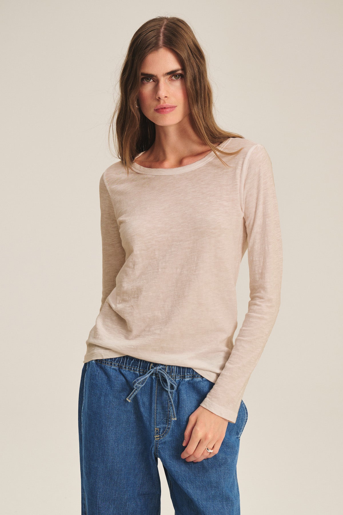 A woman wears the LIZZIE TEE by Velvet by Graham & Spencer, a soft crew neck long sleeve top in beige, paired with blue drawstring pants, standing against a plain light background.