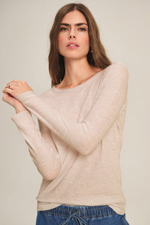 A person wears the LIZZIE TEE by Velvet by Graham & Spencer, a light beige soft crew neck long sleeve top, paired with blue drawstring pants, standing against a plain background and looking at the camera.