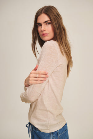 A woman with long brown hair stands sideways against a plain background, wearing the Velvet by Graham & Spencer LIZZIE TEE in beige and blue jeans, arms crossed as she looks at the camera.