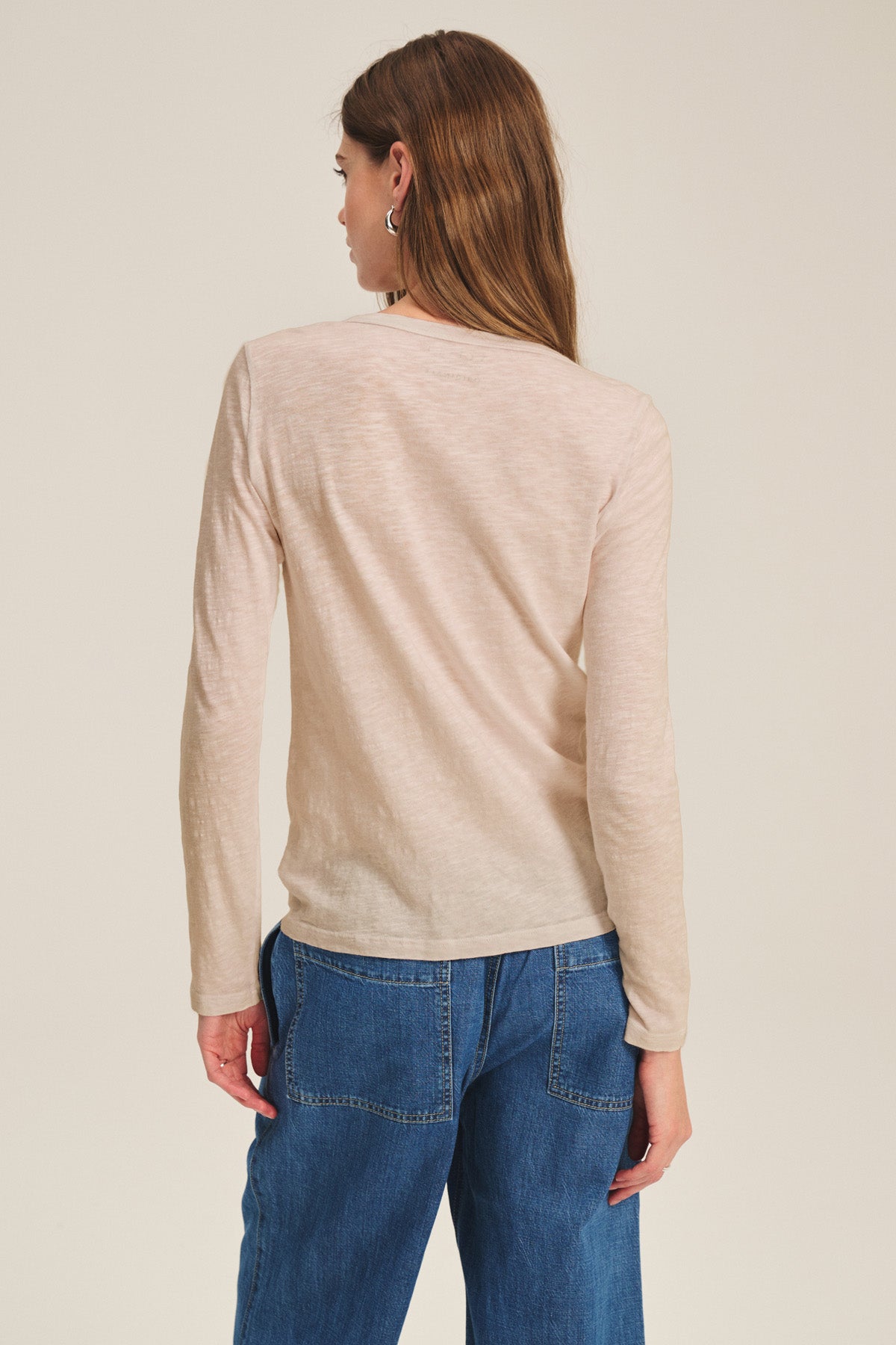 A woman with long brown hair is shown from the back, wearing the LIZZIE TEE by Velvet by Graham & Spencer—a light beige, textured cotton slub long sleeve shirt—with blue jeans against a plain background.