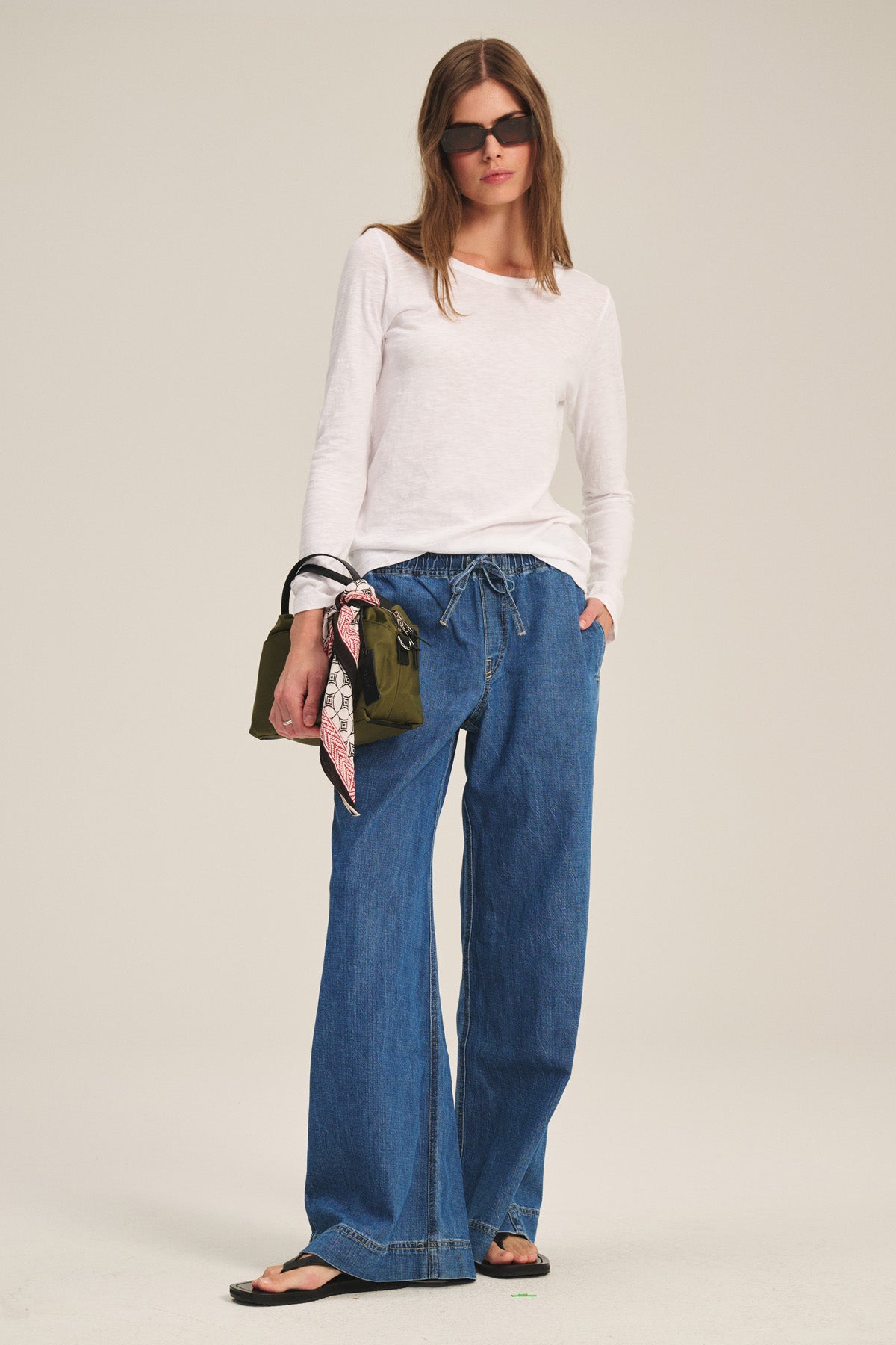 A woman in sunglasses wears the LIZZIE TEE by Velvet by Graham & Spencer, paired with loose blue jeans, black sandals, and holds an olive handbag adorned with a scarf.-40305607082177