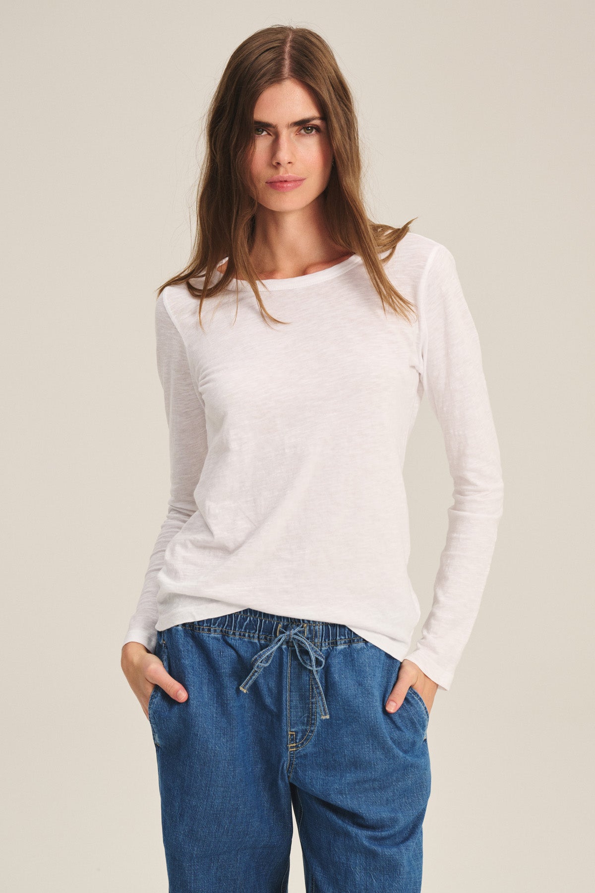 A person wears the LIZZIE TEE by Velvet by Graham & Spencer, a long-sleeved white shirt, paired with blue drawstring denim pants, standing with hands in pockets against a plain background.-40305607049409
