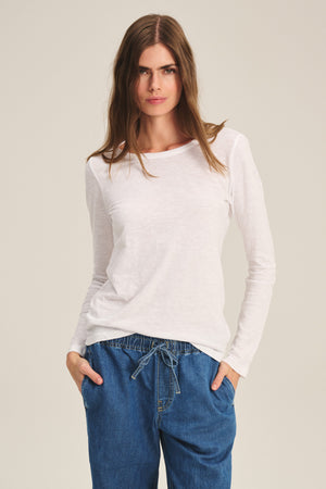 A person wears the LIZZIE TEE by Velvet by Graham & Spencer, a long-sleeved white shirt, paired with blue drawstring denim pants, standing with hands in pockets against a plain background.