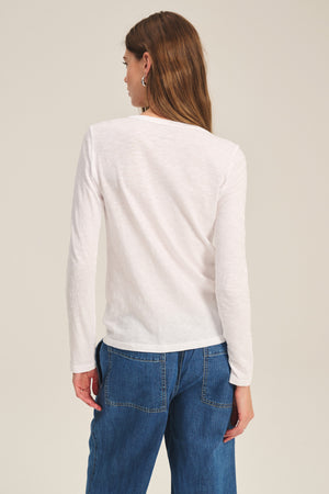 A woman faces away from the camera, wearing the LIZZIE TEE by Velvet by Graham & Spencer—a white long-sleeve shirt—paired with blue jeans, set against a plain light background.