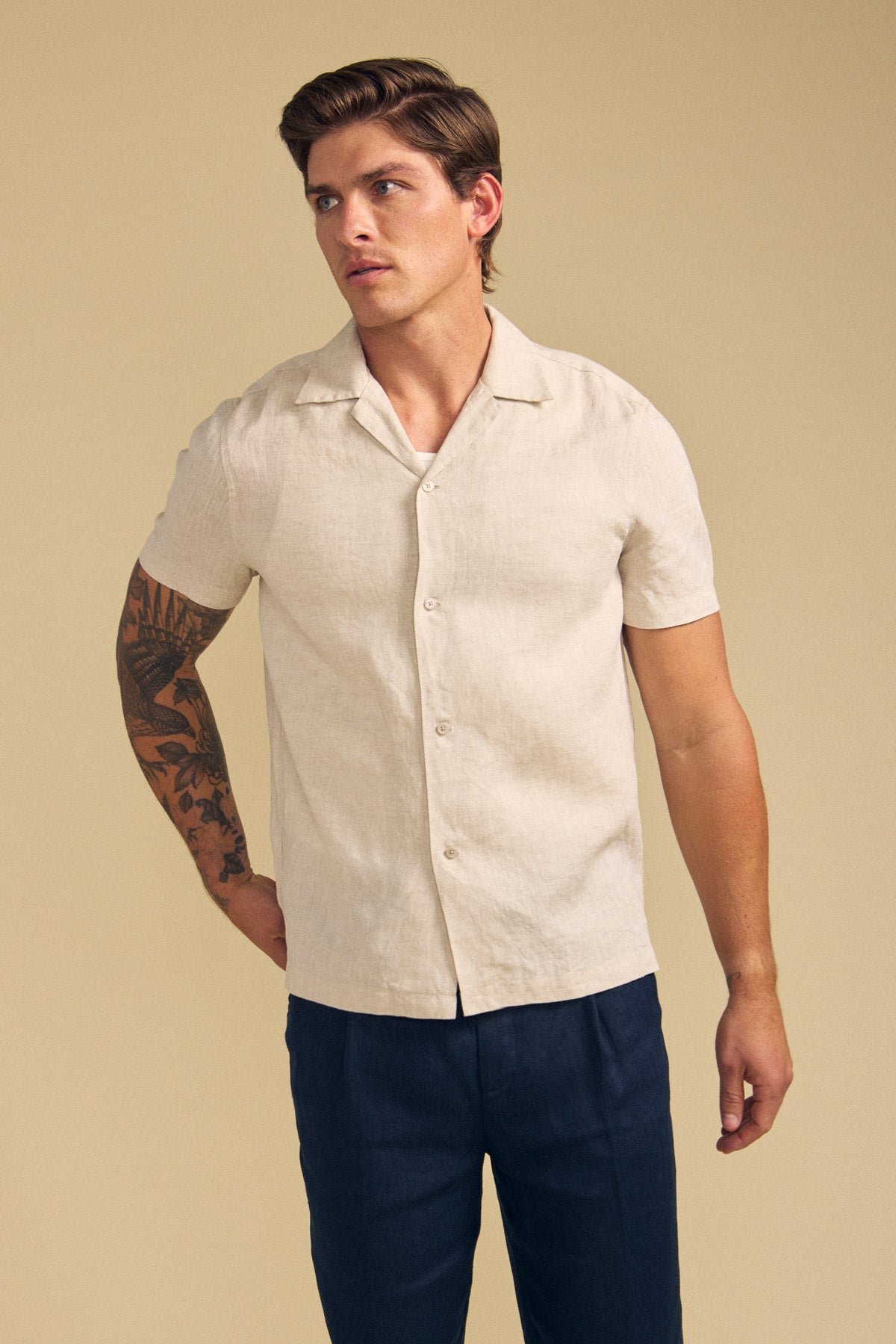 A man wears the MACKLIN LINEN SHIRT by Velvet by Graham & Spencer—a short-sleeve, light beige button-up—paired with dark trousers, standing against a plain beige background.-41702031687873