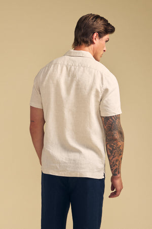 A man faces away, wearing the MACKLIN LINEN SHIRT by Velvet by Graham & Spencer—short-sleeve beige with dark pants. He has a tattoo sleeve on his right arm and stands against a plain beige background.