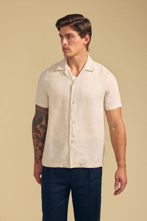 A man wears the MACKLIN LINEN SHIRT by Velvet by Graham & Spencer, a short-sleeve light beige button-up, paired with dark pants and standing against a plain beige background.