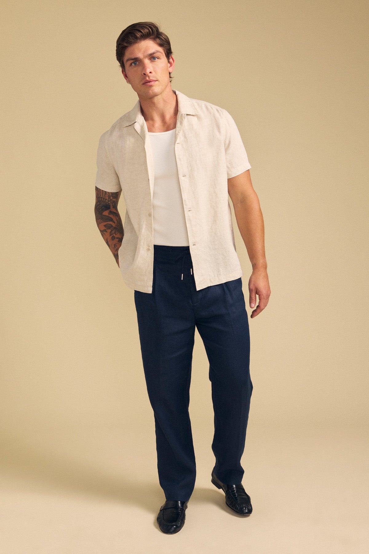 A man stands against a beige background wearing the Velvet by Graham & Spencer MACKLIN LINEN SHIRT over a white undershirt, paired with dark drawstring pants and black shoes. One of his arms features a large tattoo.