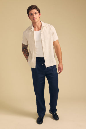 A man stands against a beige background wearing the Velvet by Graham & Spencer MACKLIN LINEN SHIRT over a white undershirt, paired with dark drawstring pants and black shoes. One of his arms features a large tattoo.