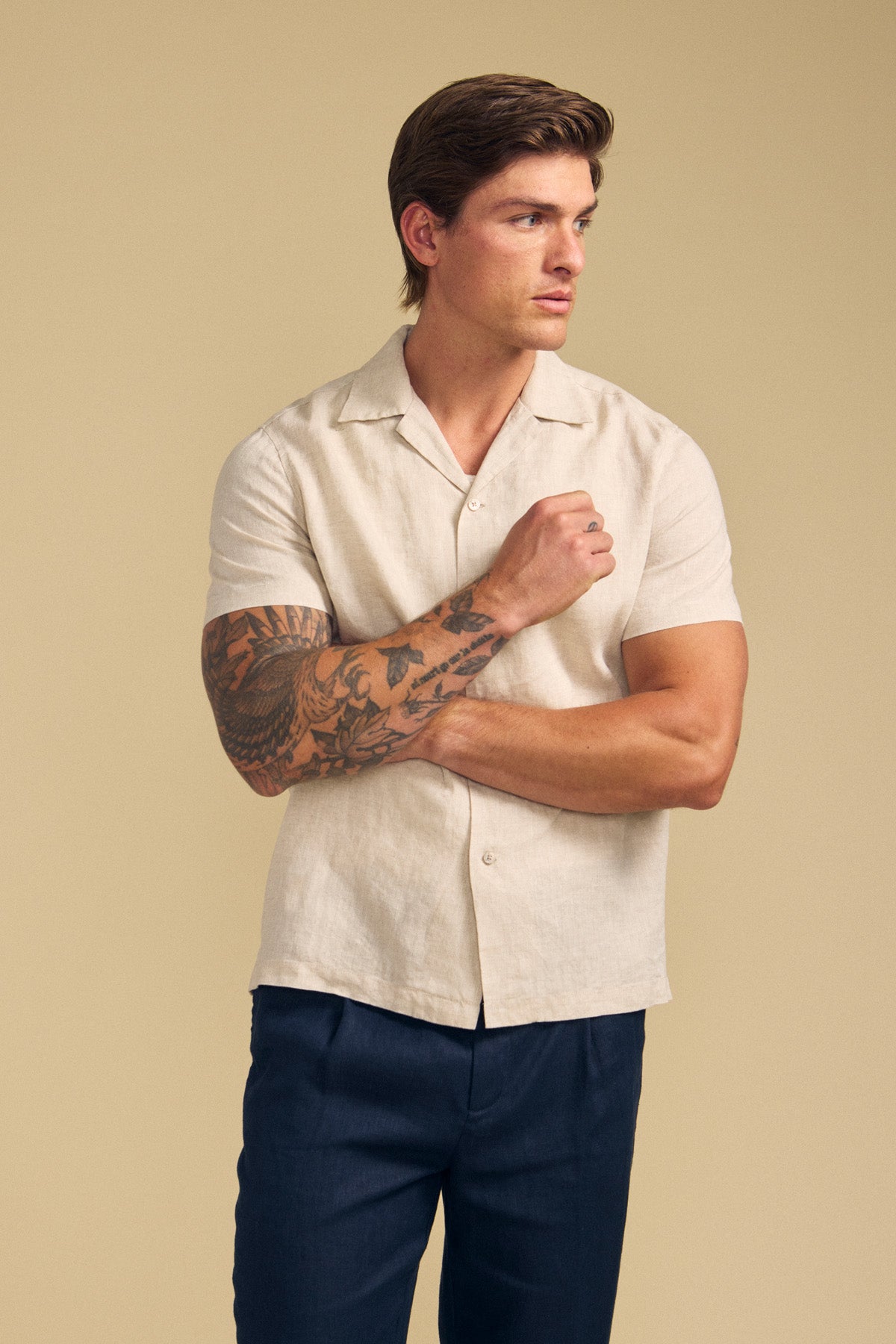 A man with short brown hair and a tattooed right arm wears the MACKLIN LINEN SHIRT by Velvet by Graham & Spencer and dark pants, standing against a plain beige background.