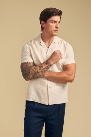 A man with short brown hair and a tattooed right arm wears the MACKLIN LINEN SHIRT by Velvet by Graham & Spencer and dark pants, standing against a plain beige background.