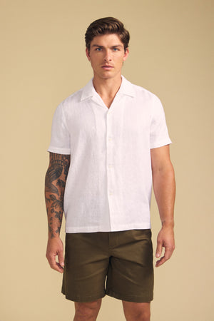 A man faces forward against a plain beige background wearing the MACKLIN LINEN SHIRT by Velvet by Graham & Spencer, a white short-sleeve button-up, paired with olive green shorts.