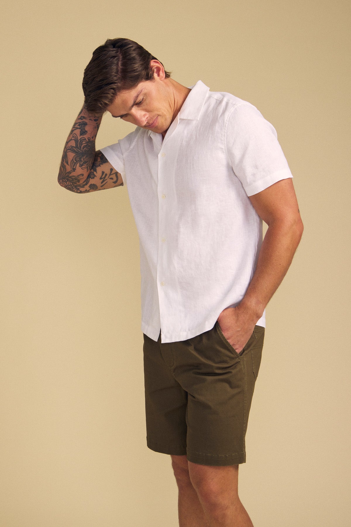 A man wears the MACKLIN LINEN SHIRT by Velvet by Graham & Spencer— a white short-sleeve button-up—paired with olive green shorts, standing against a beige background with one hand in his pocket and the other touching his head.