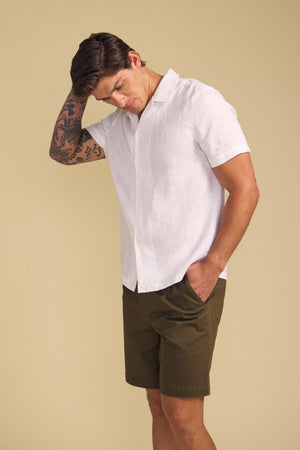 A man wears the MACKLIN LINEN SHIRT by Velvet by Graham & Spencer— a white short-sleeve button-up—paired with olive green shorts, standing against a beige background with one hand in his pocket and the other touching his head.
