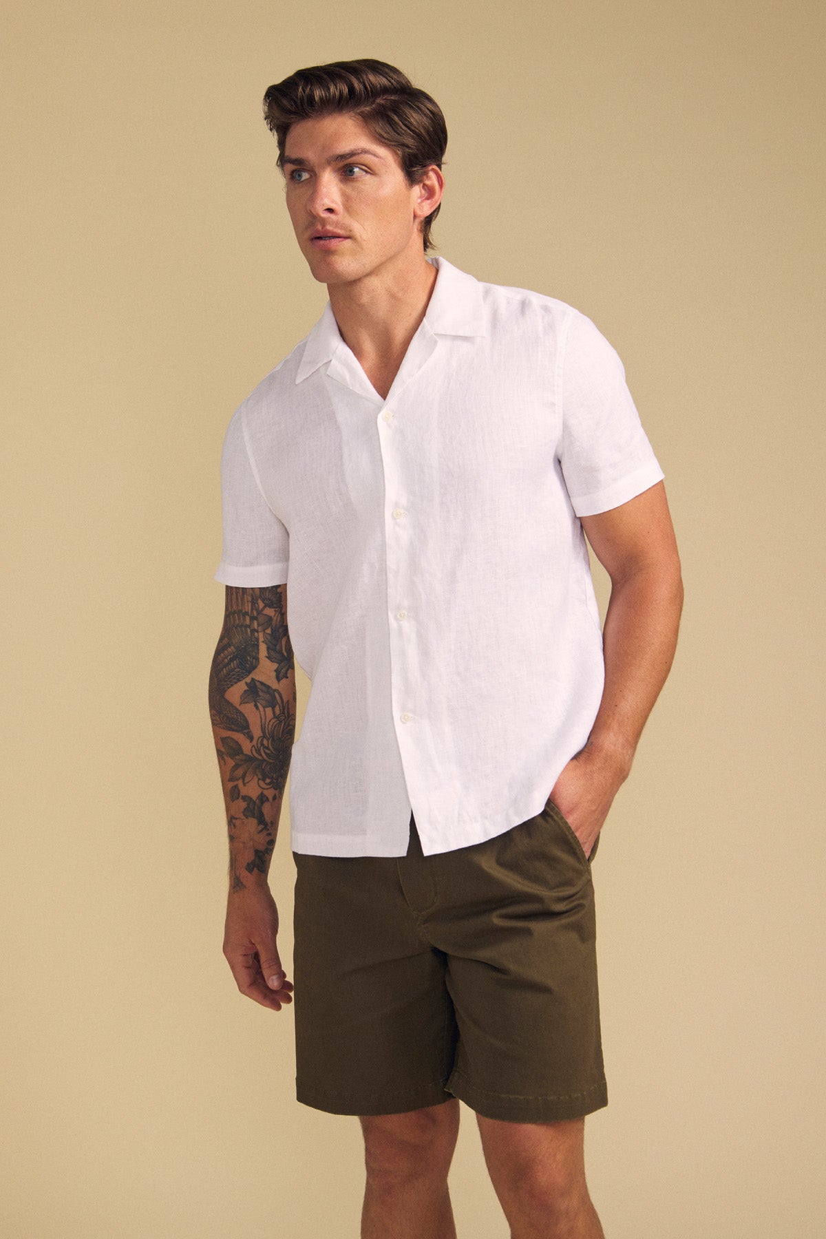 A man wears the MACKLIN LINEN SHIRT by Velvet by Graham & Spencer, a white short-sleeve button-up, paired with olive green shorts against a plain beige background.-41702031163585