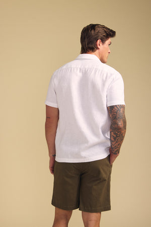 A man with a tattooed right arm wears the MACKLIN LINEN SHIRT by Velvet by Graham & Spencer, paired with olive green shorts, facing away against a plain beige background.