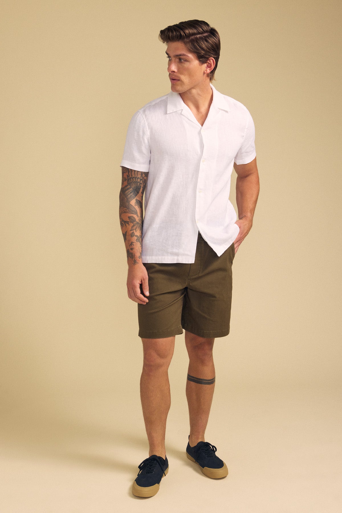 A man stands against a beige background wearing the Velvet by Graham & Spencer MACKLIN LINEN SHIRT with olive shorts and black sneakers, showcasing tattoos on his right arm and left leg.