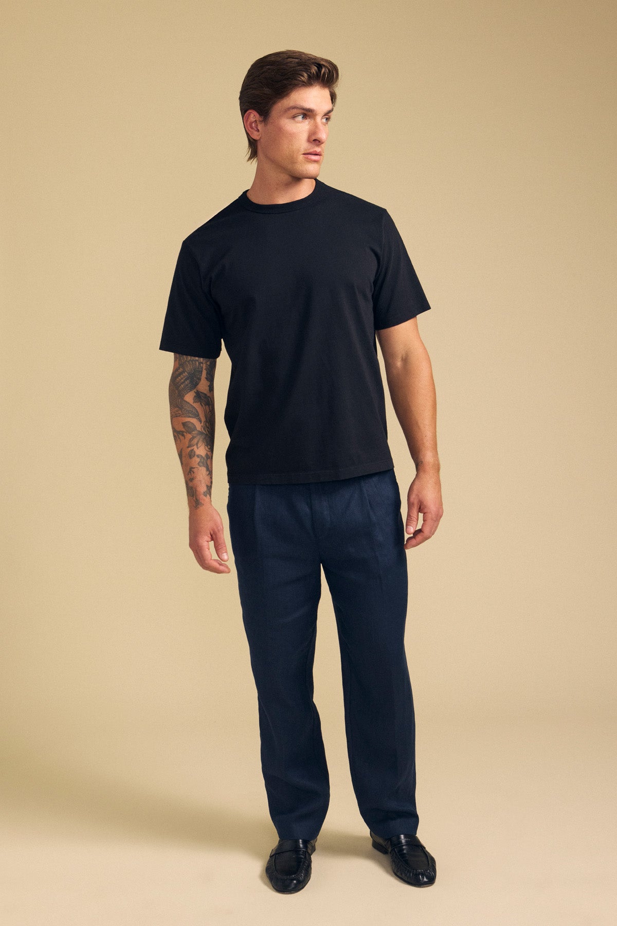 A man stands against a beige background wearing the ELLIOTT LINEN PANT from Velvet by Graham & Spencer, a black T-shirt, and black shoes, with a tattoo on his left forearm, looking to his right.
