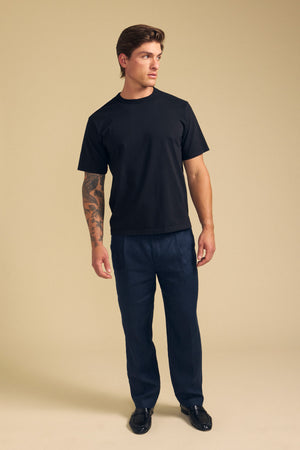 A man stands against a beige background wearing the ELLIOTT LINEN PANT from Velvet by Graham & Spencer, a black T-shirt, and black shoes, with a tattoo on his left forearm, looking to his right.
