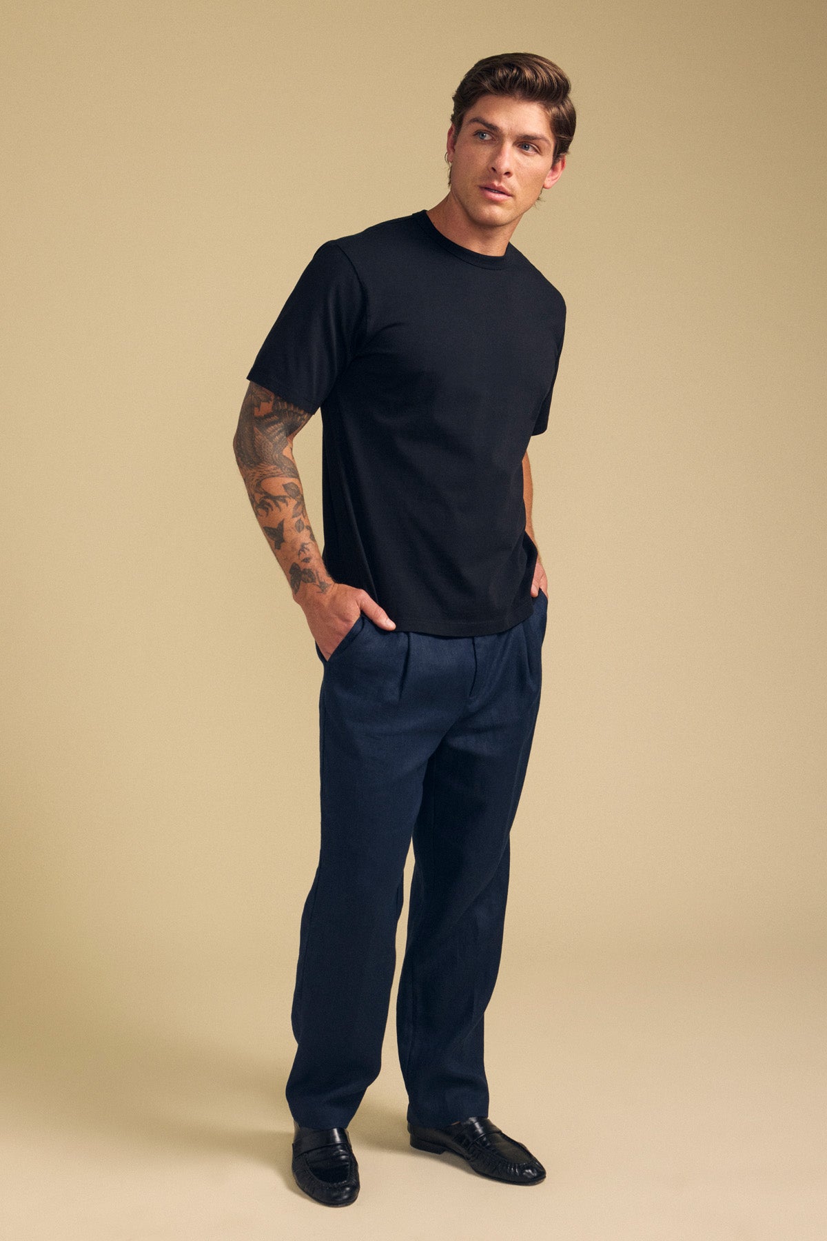 A man with tattoos wears the Velvet by Graham & Spencer ELLIOTT LINEN PANT, a plain black T-shirt, and black shoes, standing against a beige background with his hands in his pockets.-41701921915073